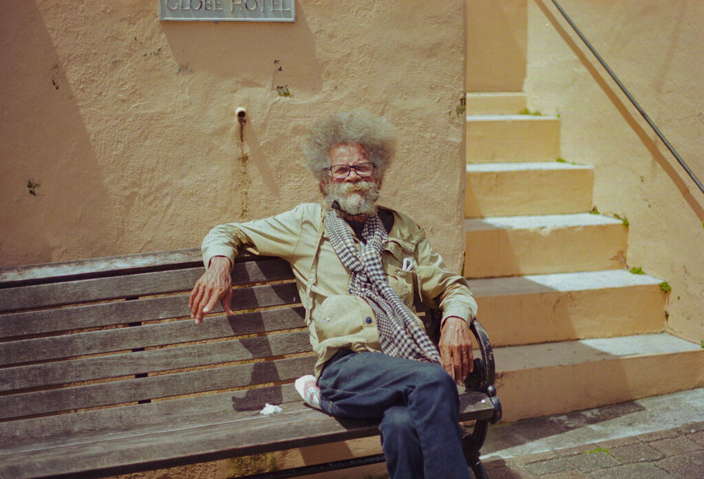 A man sitting on a park bench outside The Globe Museum in Hamilton, Bermuda. (Photo taken with subject&#39;s consent. Shot on Canon AE-1 on Kodak Portra 400 film, April 2025.)
